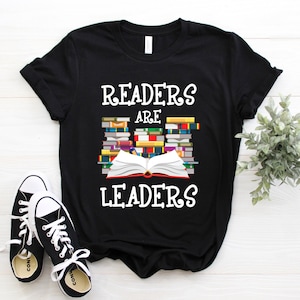 Puede incluir: Camiseta negra con el texto "Readers are Leaders" y una pila de libros con un libro abierto en la parte superior.