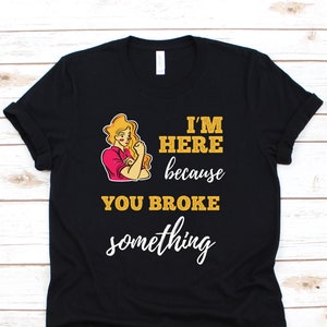 Op de afbeelding: Zwart T-shirt met een afbeelding van een vrouw met haar vuist omhoog en de tekst "Ik ben hier omdat je iets hebt gebroken."