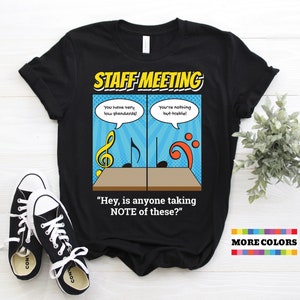 Puede incluir: Camiseta negra con un gráfico de estilo cómic de notas musicales y el texto "STAFF MEETING" y "Hey, is anyone taking NOTE of these?" El gráfico incluye el texto "You have very low standards!" y "You're nothing but treble!"
