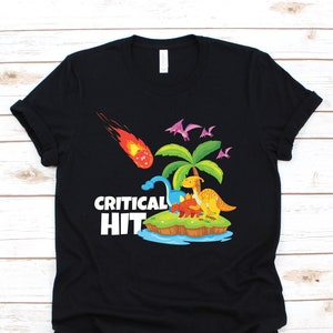 Könnte beinhalten: Schwarzes T-Shirt mit einem Aufdruck von Dinosauriern auf einer Insel mit einer Palme. Ein Meteorit fällt vom Himmel und ein Würfel ist sichtbar. Der Text "CRITICAL HIT" ist auf dem T-Shirt aufgedruckt.