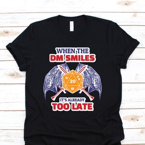 Puede incluir: Camiseta negra con un gráfico de un dado de 20 caras con espadas cruzadas y alas de murciélago. El texto "When the DM Smiles It's Already Too Late" está impreso en la camiseta.