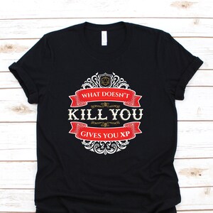 May include: Black t-shirt with a white and gold graphic design. The graphic features a red banner with the text "What Doesn't Kill You Gives You XP".
