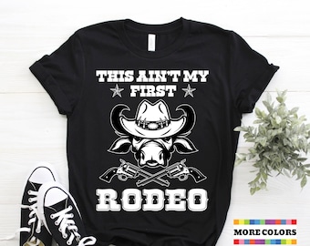 May include: Black t-shirt featuring a white graphic of a bull wearing a cowboy hat and holding two pistols. The text "This ain't my first rodeo" is printed on the shirt.