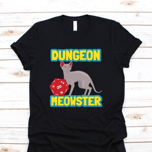 May include: Black t-shirt with a blue and yellow graphic of a cat holding a red twenty-sided die. The text "Dungeon Meowster" is printed in yellow on the shirt.