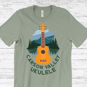 May include: Sage green t-shirt featuring a graphic of an orange ukulele with a mountain and lake scene. The text "Carson Valley Ukulele" is arched around the ukulele. The shirt is made of a soft material.