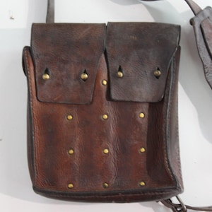 May include: A brown leather pouch with two compartments, each covered by a flap secured with a brass stud. Additional brass studs are evenly spaced on the pouch's front. A leather strap is attached for carrying.