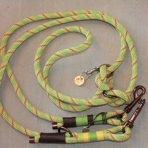 Op de afbeelding: Een groene, oranje en zwarte touwhalster voor honden met twee metalen clips en een bruine leren accent.