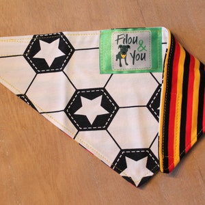 May include: A white and black soccer ball patterned bandana with a yellow border and a green label that says "Filou & You". The bandana is folded in half and has a red, yellow, and black striped fabric on the other side.