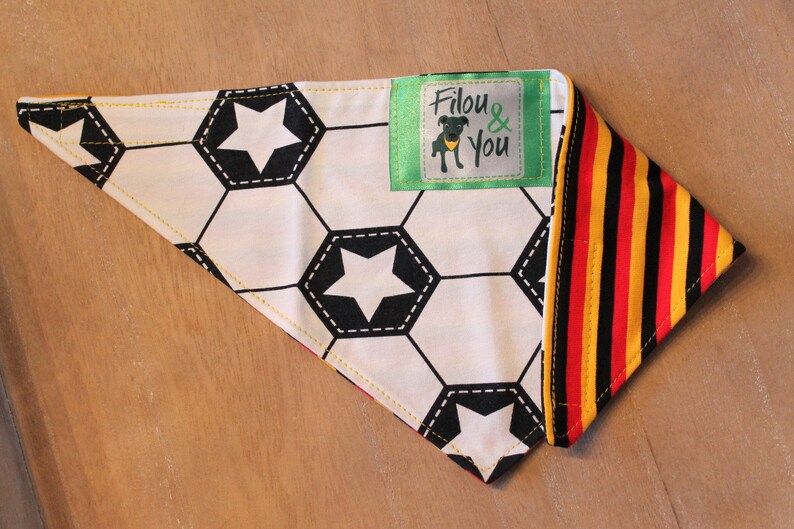 May include: A white and black soccer ball patterned bandana with a yellow border and a green label that says "Filou & You". The bandana is folded in half and has a red, yellow, and black striped fabric on the other side.