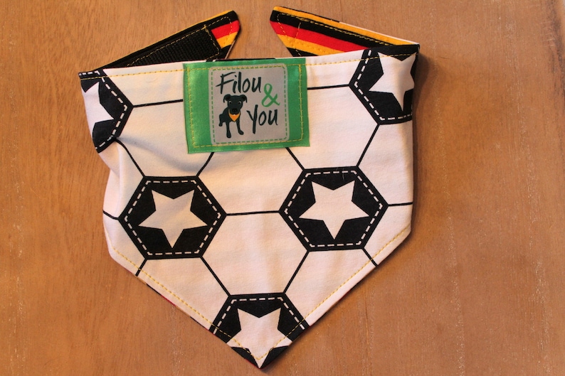 May include: A white and black soccer ball patterned dog bandana with yellow stitching. The bandana has a green label with the text "Filou & You" and a black silhouette of a dog wearing a yellow collar.