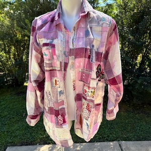 May include: A long-sleeved button-down shirt featuring a patchwork design in various shades of pink, white, and burgundy. The garment is constructed from multiple fabric squares with visible stitching, including embroidered butterfly and floral motifs.