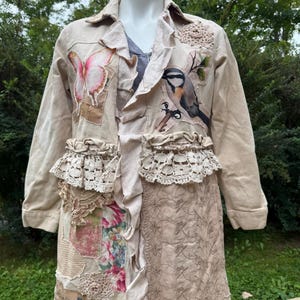 May include: A beige jacket featuring a ruffled front placket, a pink butterfly appliqué, and a bird illustration. The garment includes layered lace trim, a floral fabric patch, and an embroidered panel on the lower section with crochet accents.