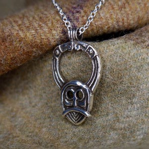 May include: A silver pendant necklace featuring a stylized face design with a beard and circular accents. The pendant hangs from a delicate chain. The background is a textured, brown fabric.