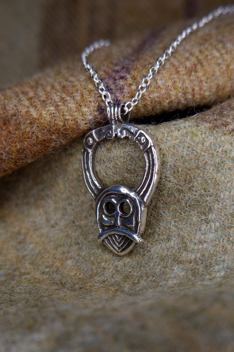 May include: A silver pendant necklace featuring a stylized face design with a beard and circular accents. The pendant hangs from a delicate chain. The background is a textured, brown fabric.