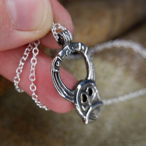 May include: A silver pendant necklace featuring a stylized, open-circle design with an owl-like face. The pendant is suspended from a delicate chain. The necklace is held in a hand against a textured, neutral-toned background.