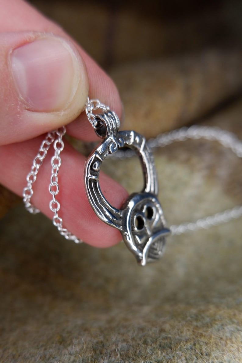 May include: A silver pendant necklace featuring a stylized, open-circle design with an owl-like face. The pendant is suspended from a delicate chain. The necklace is held in a hand against a textured, neutral-toned background.
