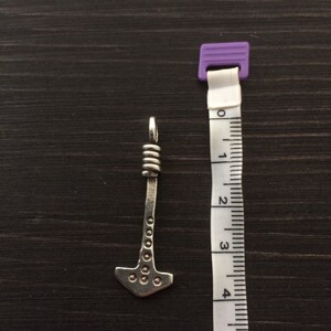 May include: A silver-colored pendant shaped like a hammer with a decorative design. The pendant is about 4 cm long and is shown next to a measuring tape.