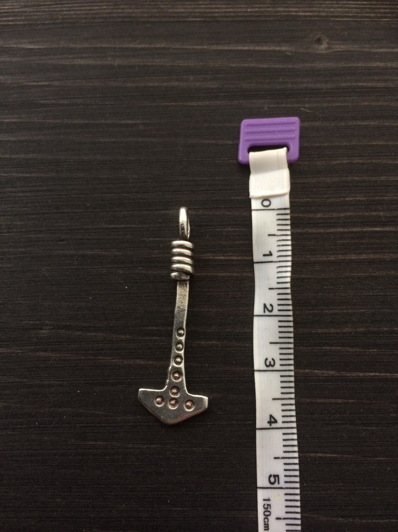 May include: A silver-colored pendant shaped like a hammer with a decorative design. The pendant is about 4 cm long and is shown next to a measuring tape.