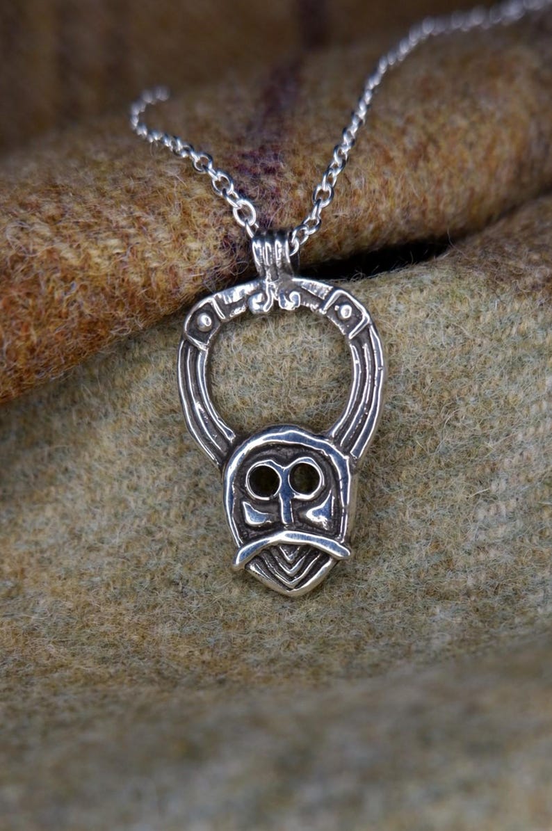 May include: A silver pendant necklace featuring a stylized Viking mask design. The pendant has a circular top and a shield-shaped bottom with a face-like design. The necklace chain is delicate and silver.