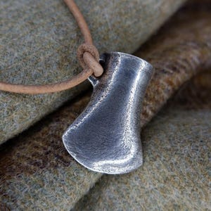 May include: A silver axe pendant on a brown leather cord. The pendant is a simple, minimalist design with a rounded head and a slightly curved blade.