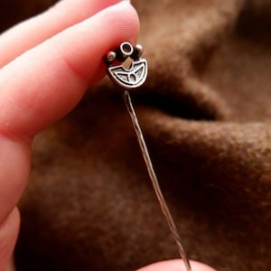 Peut inclure: Une broche en argent avec un motif décoratif. La broche est tenue dans une main et est sur un fond brun.
