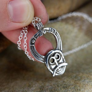 May include: A silver pendant necklace featuring a stylized Viking helmet design. The pendant is intricately detailed with curved lines and circular accents, suspended from a delicate chain. The necklace is held in a hand, showcasing the craftsmanship.