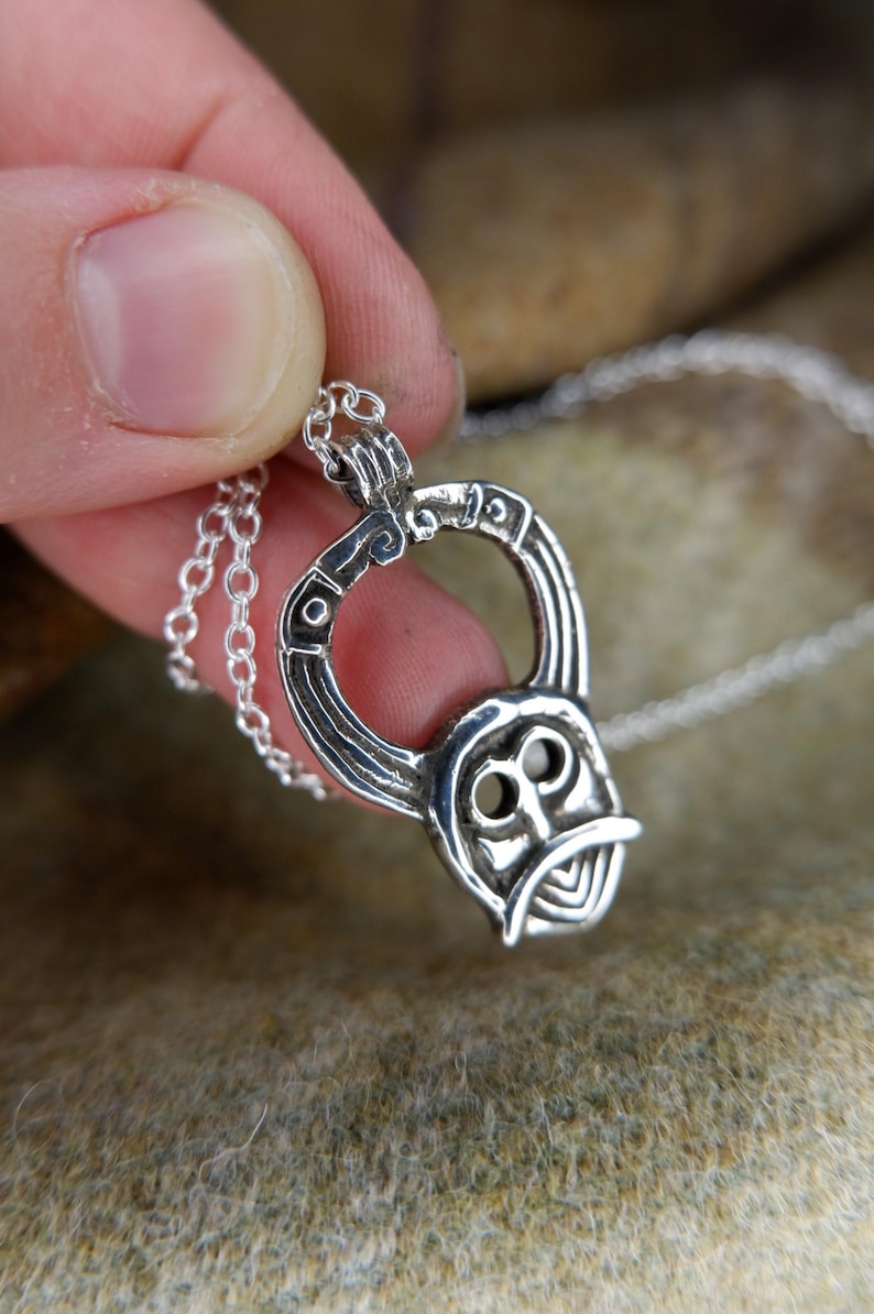 May include: A silver pendant necklace featuring a stylized Viking helmet design. The pendant is intricately detailed with curved lines and circular accents, suspended from a delicate chain. The necklace is held in a hand, showcasing the craftsmanship.