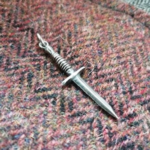 Puede incluir: Un colgante de daga de plata con un mango retorcido. El colgante está sobre una superficie peluda marrón y rosa.