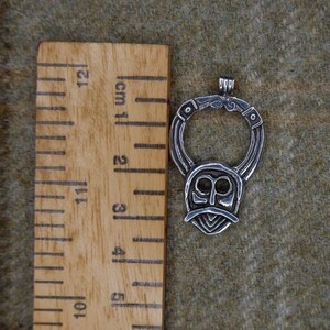 May include: Silver-toned pendant with a stylized face design, approximately 2 inches in height. The pendant features a circular top with intricate detailing and a face-like shape below. The pendant is displayed on a textured, olive-green surface.