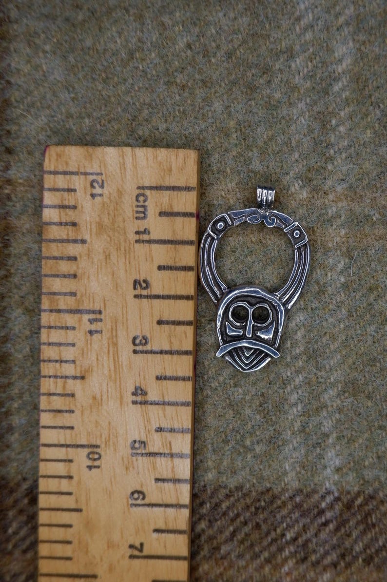 May include: Silver-toned pendant with a stylized face design, approximately 2 inches in height. The pendant features a circular top with intricate detailing and a face-like shape below. The pendant is displayed on a textured, olive-green surface.