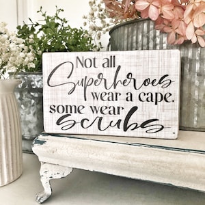 May include: White wooden sign with black text that reads "Not all superheroes wear a cape. Some wear scrubs."