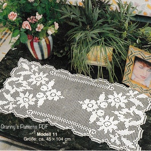 May include: White crocheted table runner with a floral design. The runner is on a black table with a vase of pink roses and a framed photo. The text on the runner reads "Granny's Patterns PDF Modell 11 Größe: ca. 45 x 104 cm".