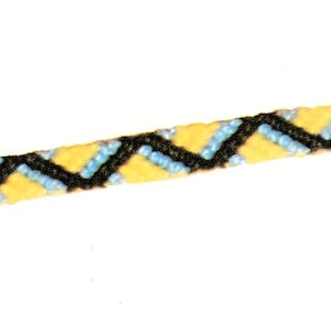 Könnte beinhalten: Ein handgefertigtes Freundschaftsarmband mit Zickzackmuster. Das Armband besteht aus gelben, hellblauen und schwarzen Fäden. Das Armband wird vor einem weißen Hintergrund präsentiert, um seine Farben und sein kompliziertes Gewebe hervorzuheben.