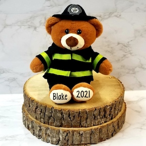 May include: Brown teddy bear dressed as a firefighter with a black hat and yellow reflective tape. The bear is sitting on a wooden platform and has the names "Blake" and "2021" written on its feet.