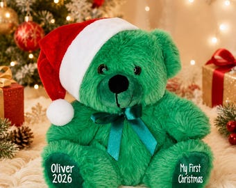 May include: A plush green teddy bear wearing a red and white Santa hat and a dark green satin ribbon around its neck. The bear's left foot is embroidered with the text Oliver 2026, and the right foot features the text My First Christmas.
