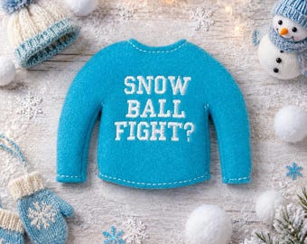 May include: A bright blue felt sweater ornament featuring the white embroidered text SNOW BALL FIGHT? surrounded by a knit hat, a snowman, and blue knit mittens with snowflake patterns on a textured white surface with scattered snowballs.