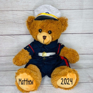 May include: A brown teddy bear wearing a navy blue uniform with a white belt and a white hat with a yellow band. The bear's paws are embroidered with the name "Matthew" and the year "2024".
