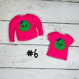 May include: Two pink felt elf shirts with a green smiling cloud design. The larger shirt has long sleeves and the smaller shirt has short sleeves. Both shirts have a purple bow on the cloud. The text #6 is visible below the shirts.