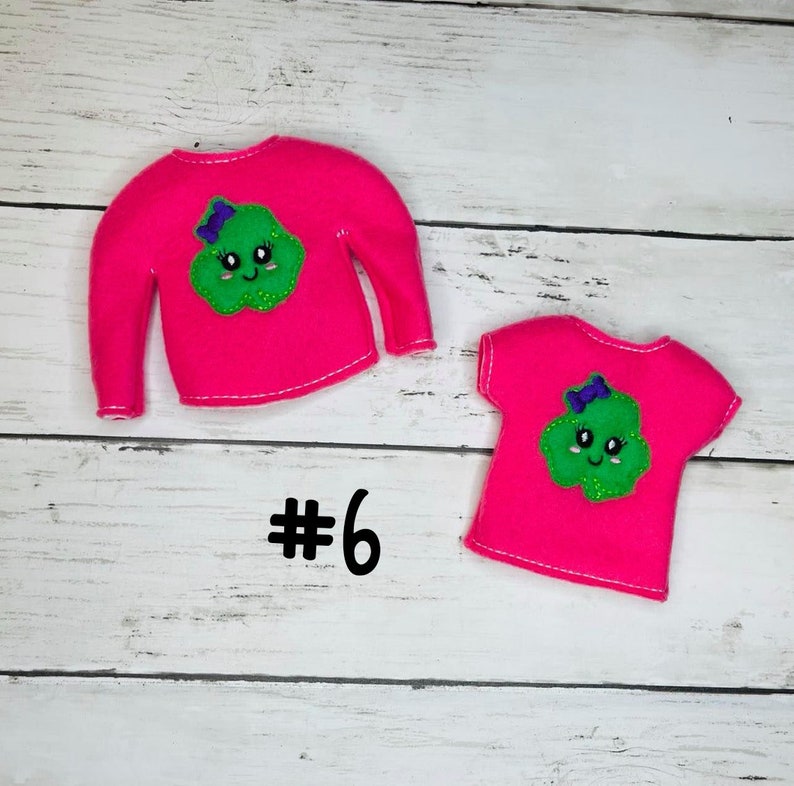 May include: Two pink felt elf shirts with a green smiling cloud design. The larger shirt has long sleeves and the smaller shirt has short sleeves. Both shirts have a purple bow on the cloud. The text #6 is visible below the shirts.