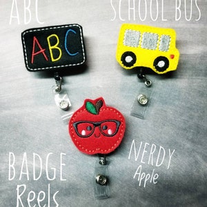 May include: Three badge reels with school-themed designs. One is a black rectangle with "ABC" in colorful letters, another is a yellow school bus, and the last is a red apple wearing glasses. The words "Badge Reels" and "Nerdy Apple" are also visible.