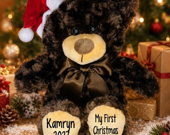 May include: A dark brown plush teddy bear wearing a red and white Santa hat and a brown satin ribbon bow around its neck. The bear's light tan foot pads feature black text that reads Kamryn 2027 on the left and My First Christmas on the right.