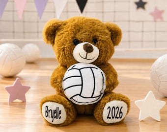 May include: A brown plush teddy bear with a white muzzle and dark eyes, holding a white volleyball with black lines. The bear has white foot pads, with the name Bryelle printed in black on the left foot and the year 2026 on the right foot.