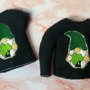 May include: Two black elf shirts with a green and white gnome holding a shamrock embroidered on the front. One shirt has short sleeves and the other has long sleeves.