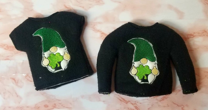 May include: Two black elf shirts with a green and white gnome holding a shamrock embroidered on the front. One shirt has short sleeves and the other has long sleeves.
