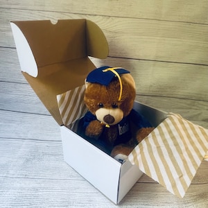 May include: A brown teddy bear wearing a blue graduation cap and gown is sitting inside a white gift box with a brown and white striped tissue paper. The bear has a yellow tassel on its cap and the name "Isaiah" is embroidered on its gown.
