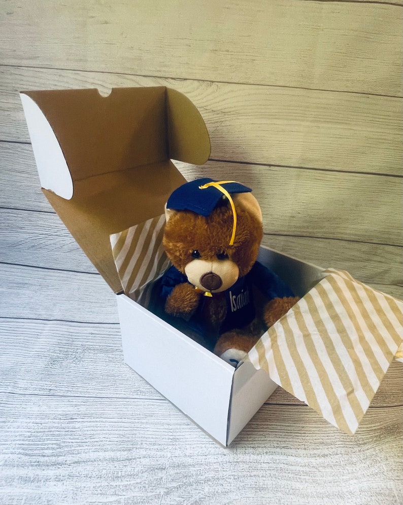 May include: A brown teddy bear wearing a blue graduation cap and gown is sitting inside a white gift box with a brown and white striped tissue paper. The bear has a yellow tassel on its cap and the name "Isaiah" is embroidered on its gown.
