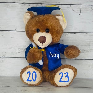 May include: A brown teddy bear wearing a blue graduation gown and cap. The bear is holding a white scroll in its right paw. The gown has a gold trim and the name "Avery" is embroidered on the front. The bear's paws have the numbers "20" and "23" written on them.