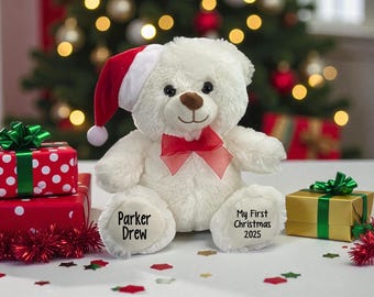 May include: A white teddy bear wearing a Santa hat and a red bow. The bear has the text "Parker Drew" on one foot and "My First Christmas 2025" on the other. Christmas-themed gifts and a decorated tree are in the background.