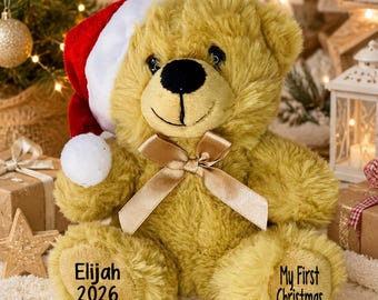 May include: A golden-brown plush teddy bear wearing a red and white Santa hat and a gold satin ribbon around its neck. The bear's left foot is embroidered with the text Elijah 2026 and the right foot with the text My First Christmas.