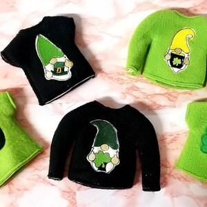 May include: Five felt elf shirts with different St. Patrick's Day designs.  Two shirts are black with a green leprechaun design. Two shirts are green with a gold pot of gold and shamrock design. One shirt is green with the word "LUCK" and a shamrock design.